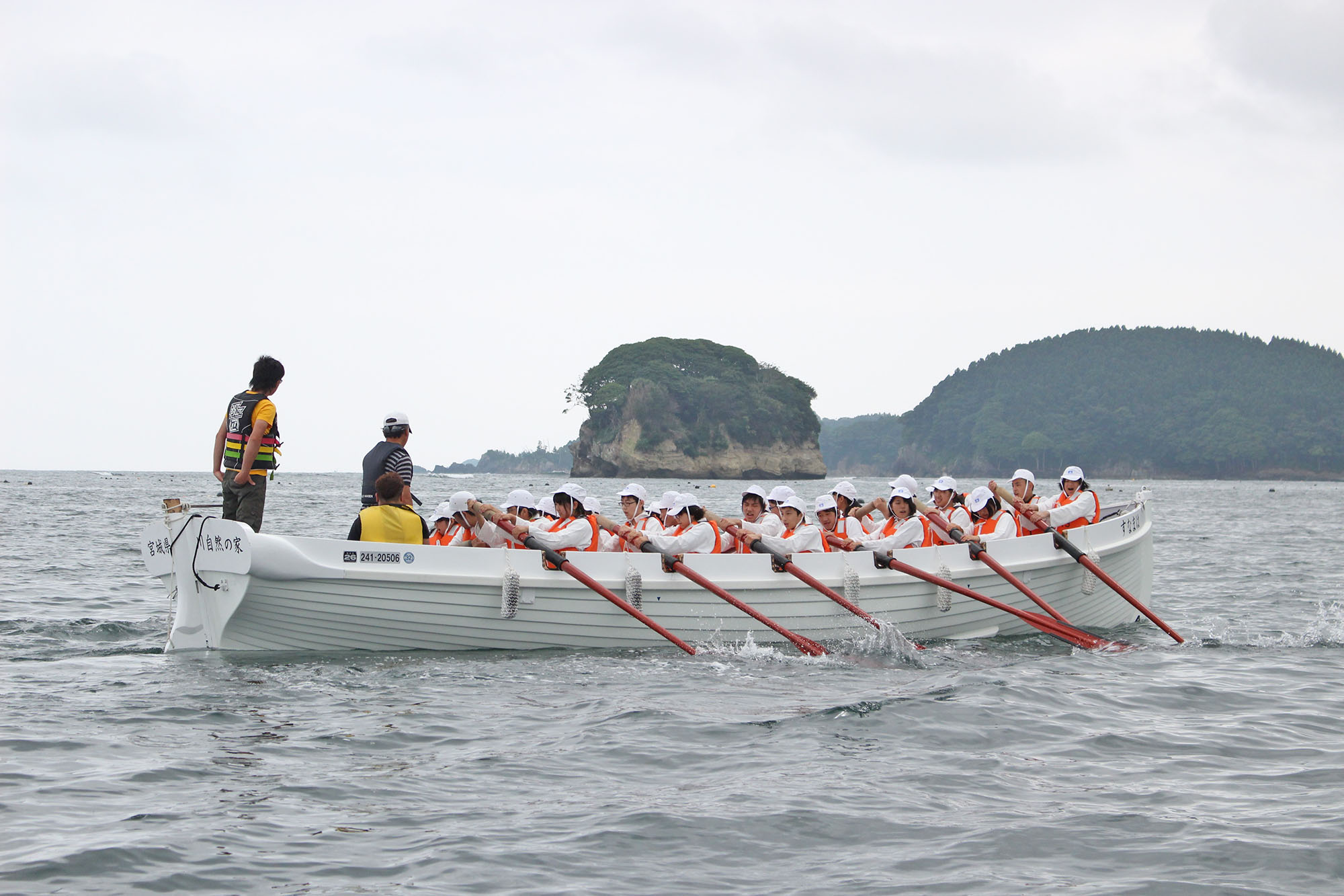 大きな手こぎボートに乗った多くの若者たちがオールを揃えて漕ぎ、指導者らしき大人が見守る中、穏やかな海の上で力を合わせて進んでいる様子を捉えた写真
