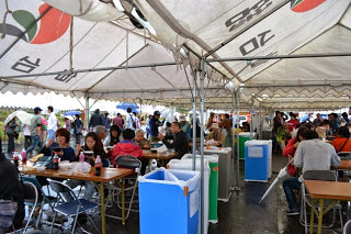 テントの下にたくさんのテーブルと椅子が並べられ、訪れた人々が食事や会話を楽しみながら思い思いに過ごしている様子を写した写真