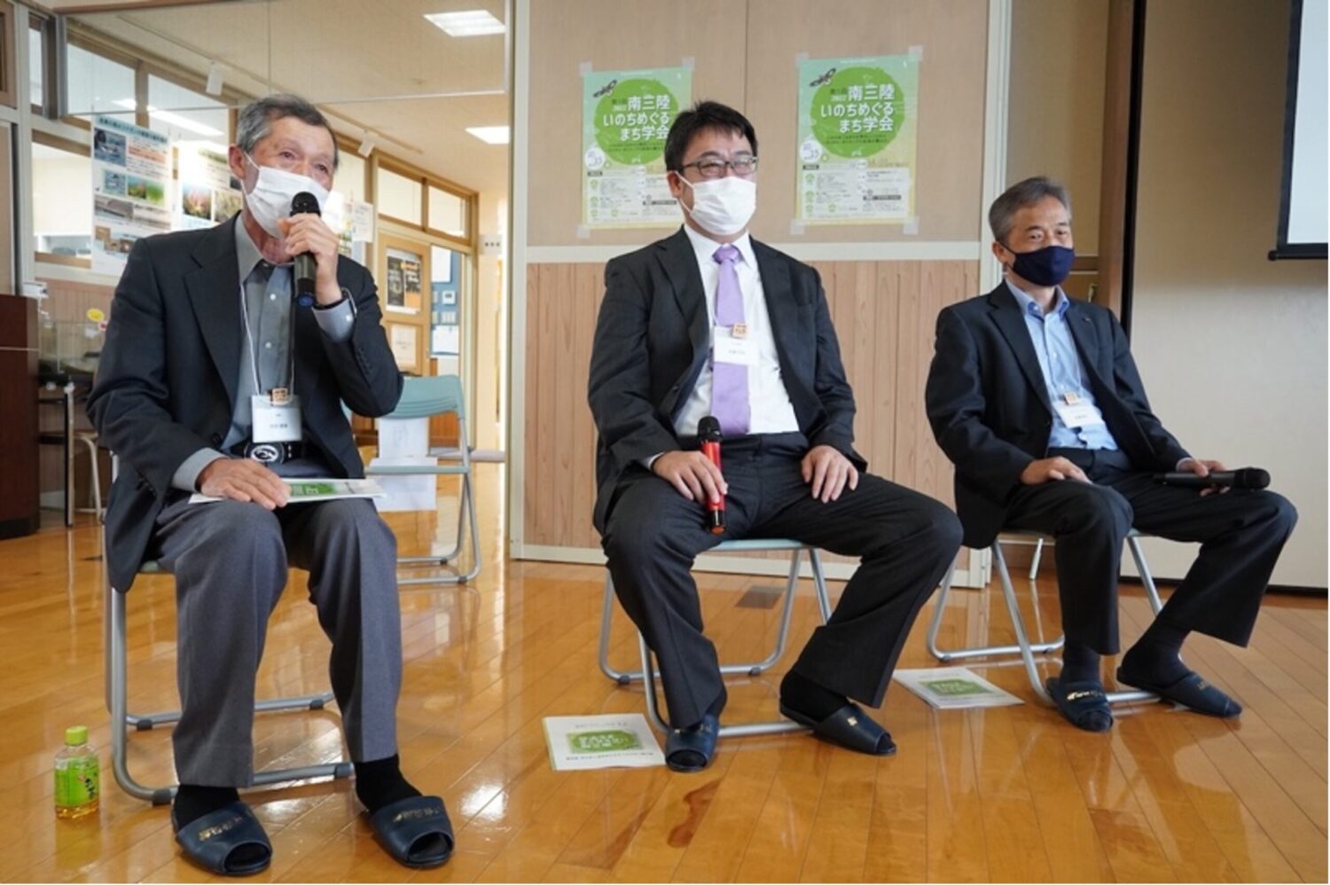 スーツ姿の関係者が3人椅子に座っており、左の阿部勝善さんがマイクを持ち話をする様子の写真