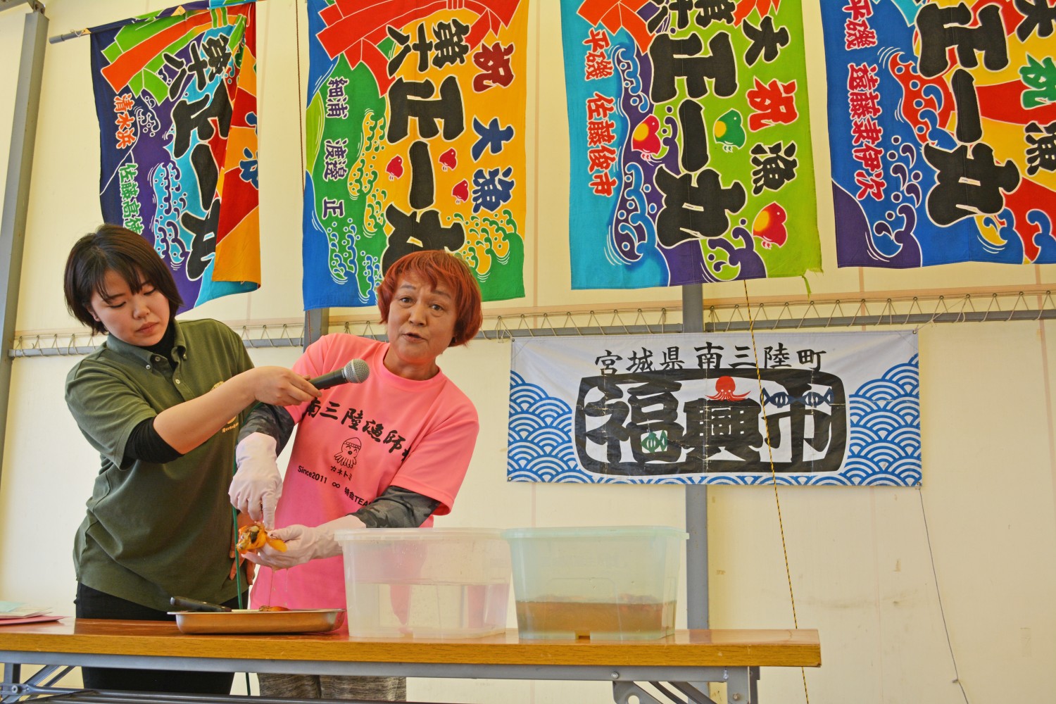 カラフルな大漁旗が飾られた福興市のステージ上で、緑色のシャツを着た女性がマイクを向けるなか、ピンクのTシャツを着た女性が水槽から取り出したホヤを持ちながら剥き方を説明している写真