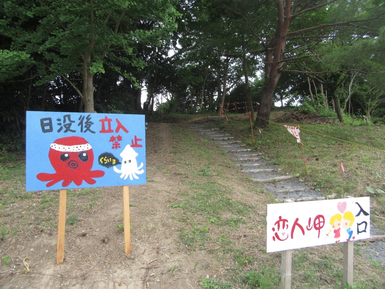木々に囲まれた小さな丘へ続く道の手前に2枚の看板が立っており、左の青い看板には赤いタコと白いイカのイラストとともに「日没後立入禁止」「くらいヨ」と書かれ、右の看板には手をつないだ2人の人物とハートのイラストが描かれ「恋人岬入口」と書かれている写真