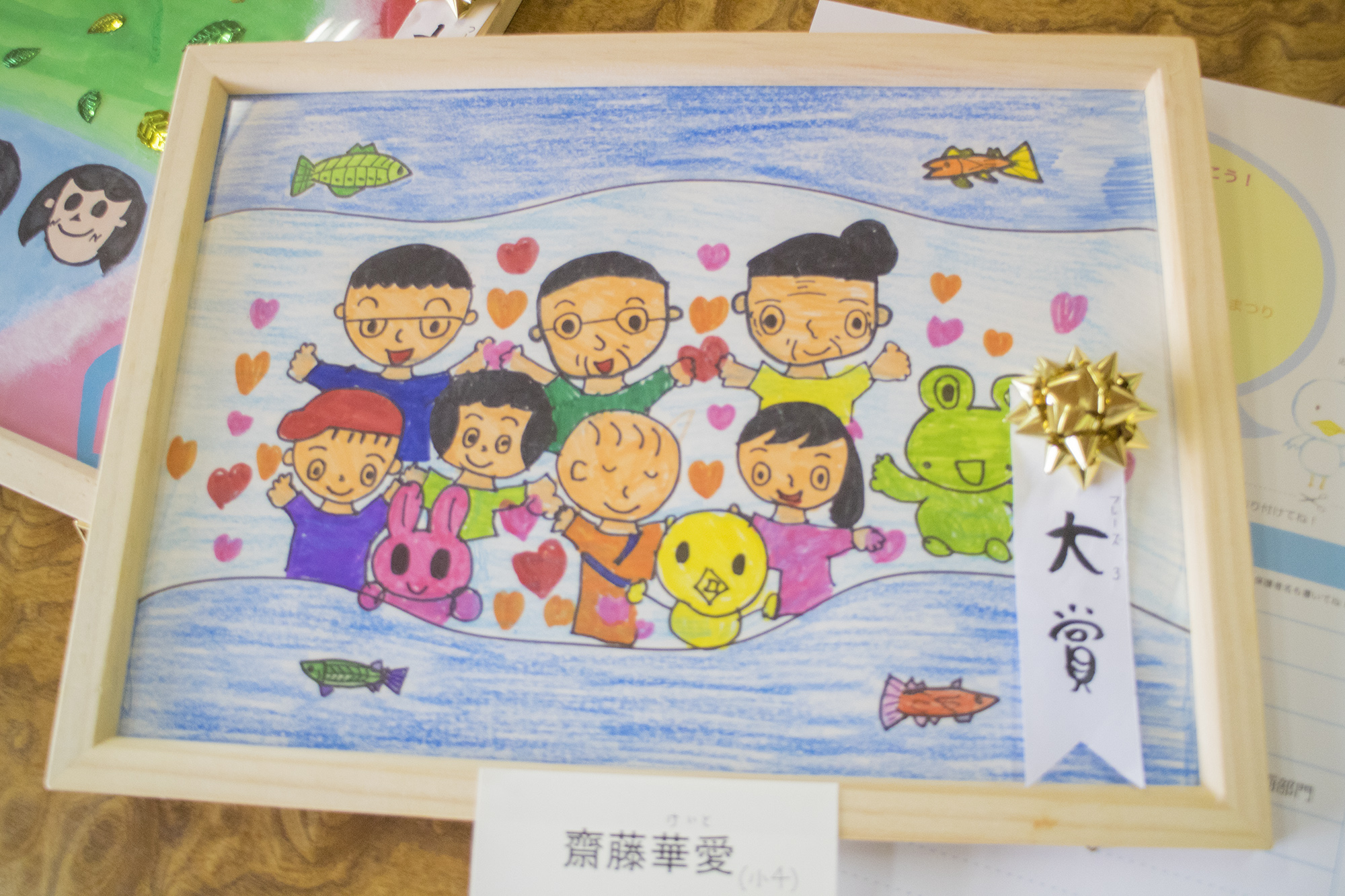 「大賞」と書かれたリボンが貼られたカラフルな受賞作品の絵には、川の中で手を上げている複数の人々と動物のキャラクターたちが笑顔で描かれている写真