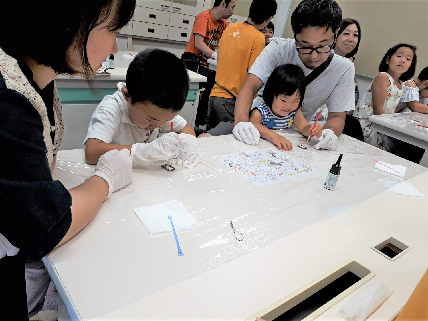 小さな子どもたちが白い手袋をつけて、親と一緒に筆で何かを塗ったりしてストラップを作っている写真