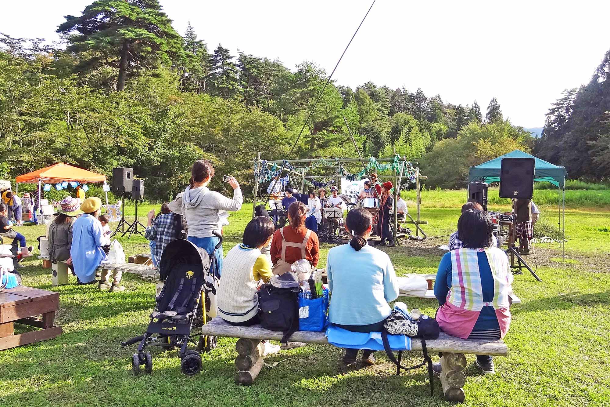 木々に囲まれた芝生の広場で、木のベンチに座った人たちが手作り感のあるステージで演奏を楽しんでいる音楽ライブの写真