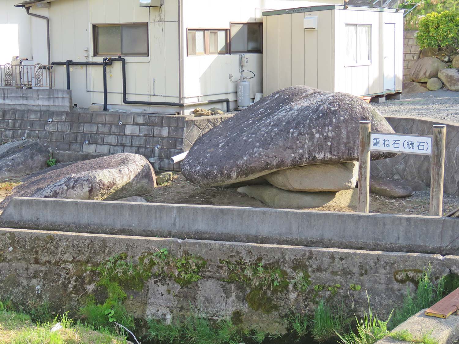 民家の傍にある重ねられた巨大な岩の近くに「重ね石（続石）」と書かれた看板が設置されており、不思議な形の石が目立つ写真