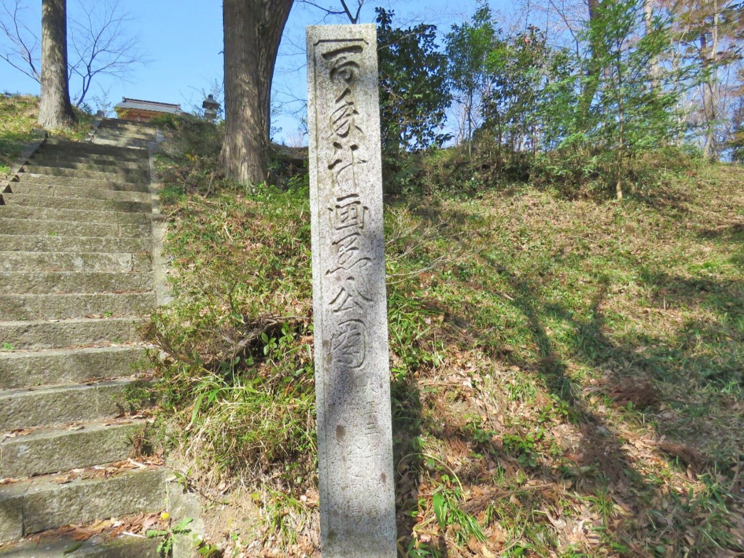 背景に木立が茂る石段の脇に、文字が刻まれた石碑が立っている写真