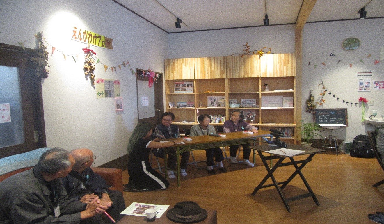 3人の高齢者の女性がテーブルに座り、女性スタッフが前方でマイクを持って説明をしながらクイズを進行している写真