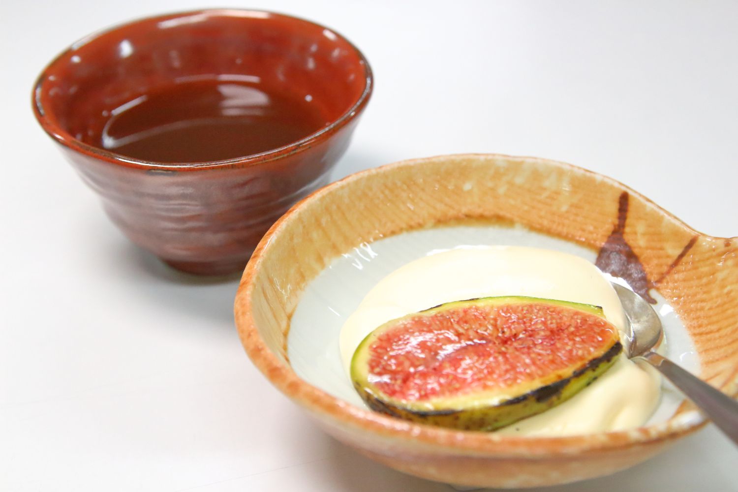 ベージュ色のお皿に白いクリームとスライスされ縁に焦げ目がついた無花果が盛り付けられ、朱色のお猪口に液体が注がれた「焦がしいちじくと梅クリーム」のデザートの写真