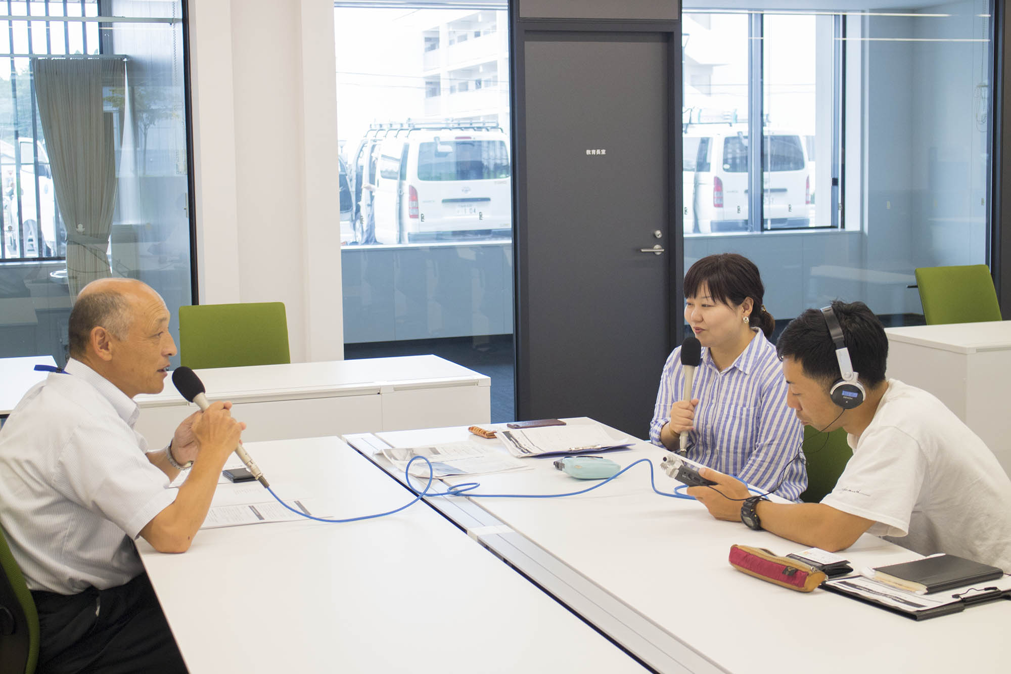 リポーターの女性とヘッドホンをして機材を持った男性が会議室のテーブルで向かい合って座った三浦さんにインタビューをしている写真