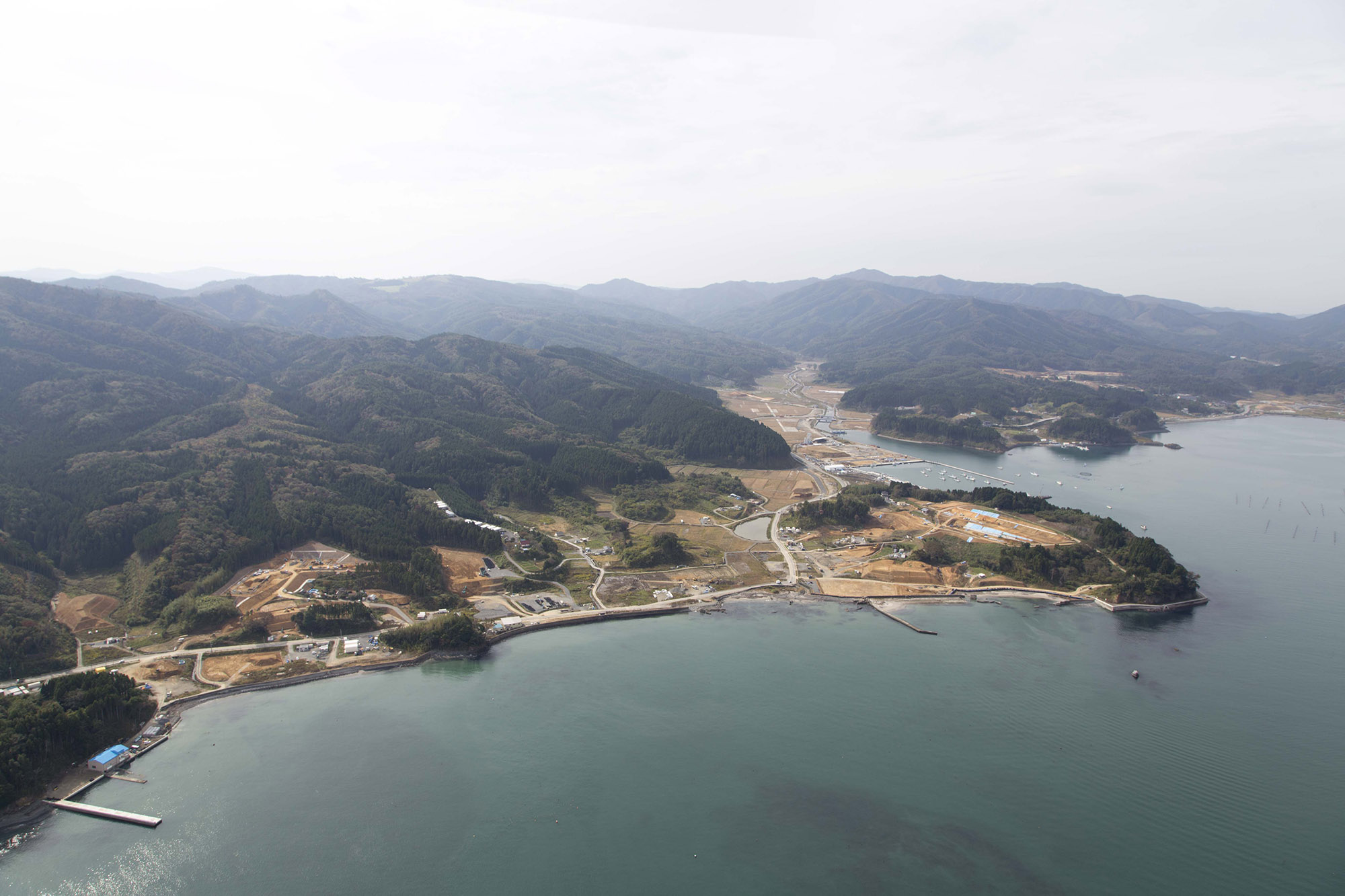 穏やかな海に面した南三陸町の海岸沿いの町並みと、その背後に広がる広大な山々や森林が映っており、港や防潮堤、建設中の建物、田畑、そして曲がりくねった道路が見える、山々に囲まれた入り組んだ湾を上空から撮影した南三陸町の写真