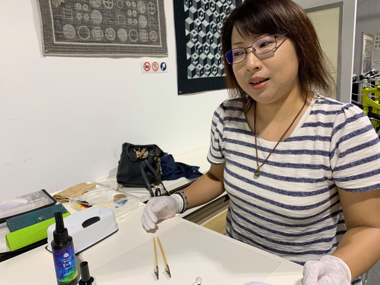 ボーダー柄のシャツを着た女性が白い手袋をはめ、作業台の上に青い液体の入ったボトルに道具や素材を広げて座っている写真