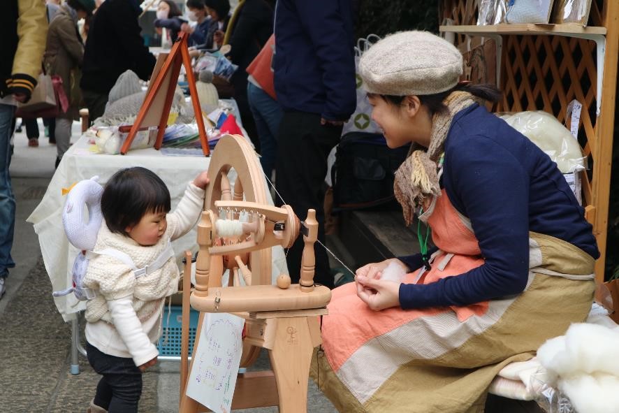 小さな女の子が糸車に興味を持って回そうとしており、店番の若い女性が微笑みながらその様子を見ている糸つむぎ体験のブースの写真