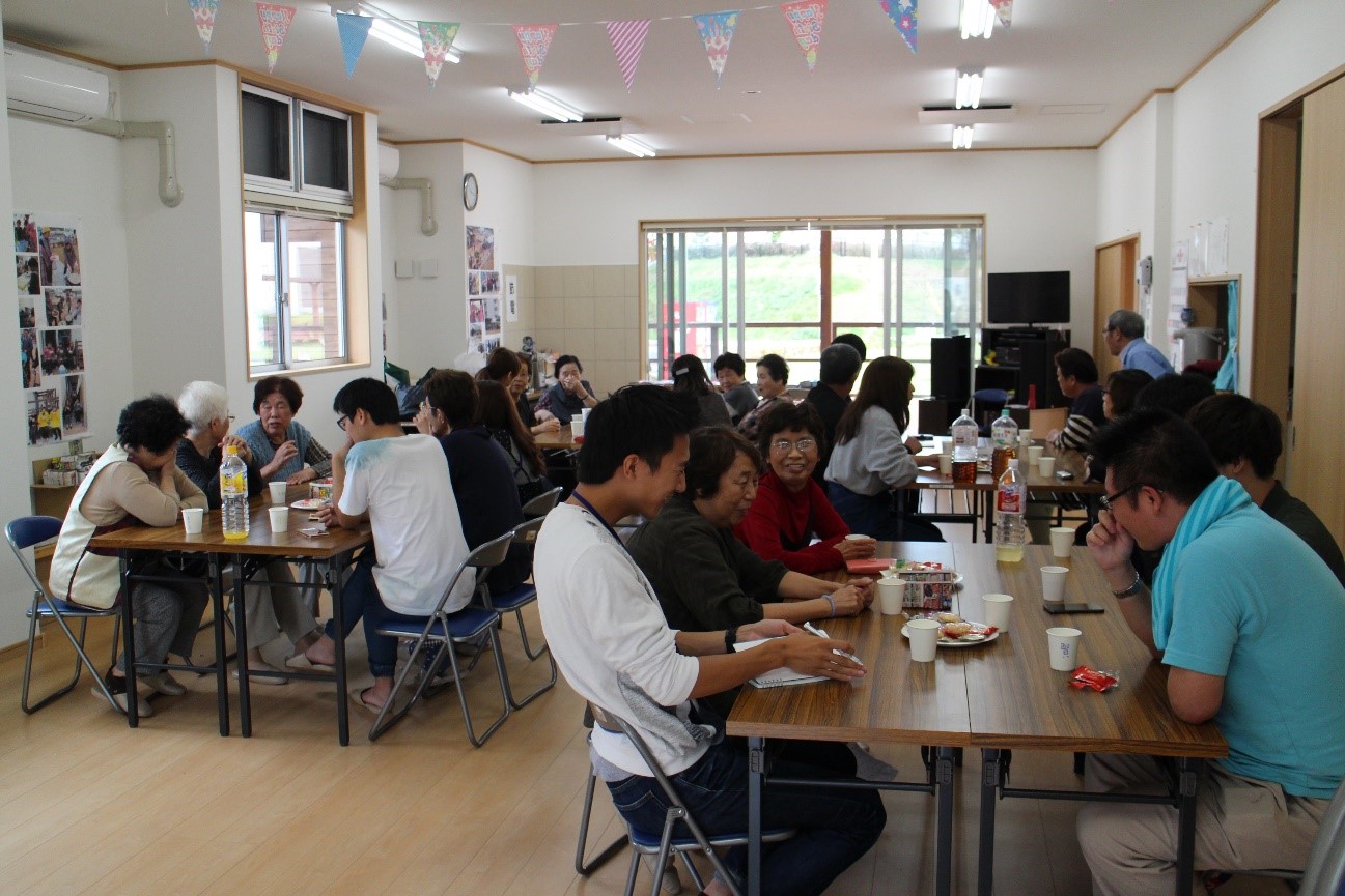 部屋の中で、複数の人々がテーブルに座り、談笑しながら食事や飲み物を楽しんでいる様子が写っている写真