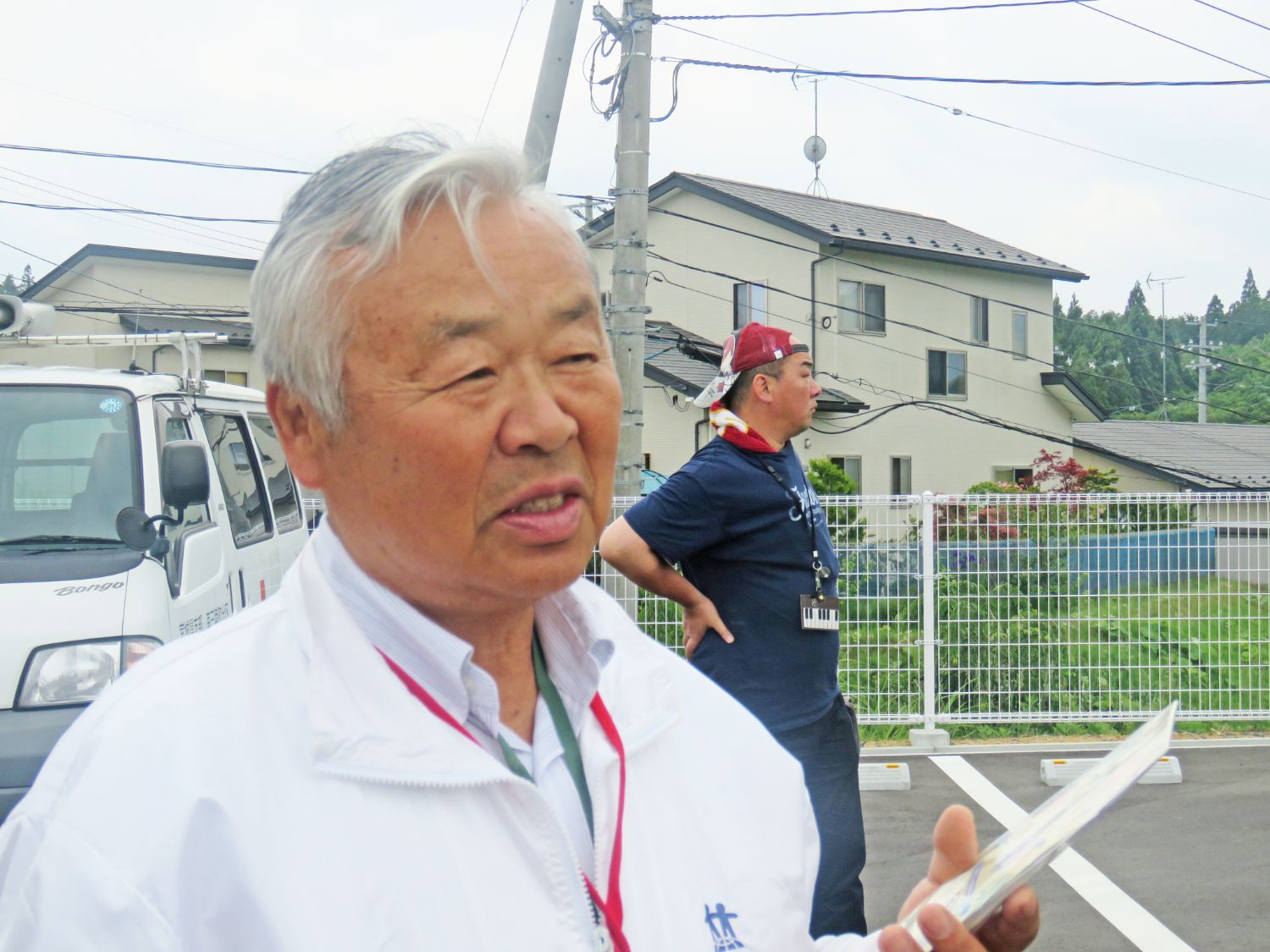 背景には住宅見え、車が停まっている駐車場で、白いジャンパーを着た白髪の高齢男性が話している様子をアップで撮影した写真