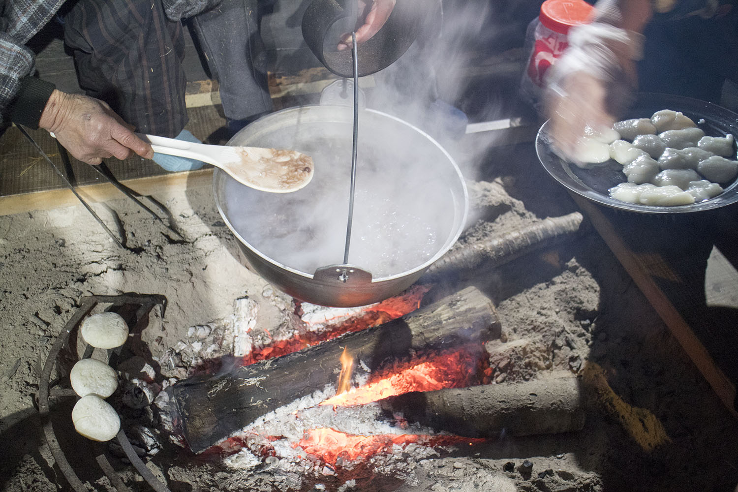 囲炉裏にまきが燃える中、大鍋で小豆が煮られており、傍らでお餅が焼かれ、しゃもじを使って調理している様子の写真