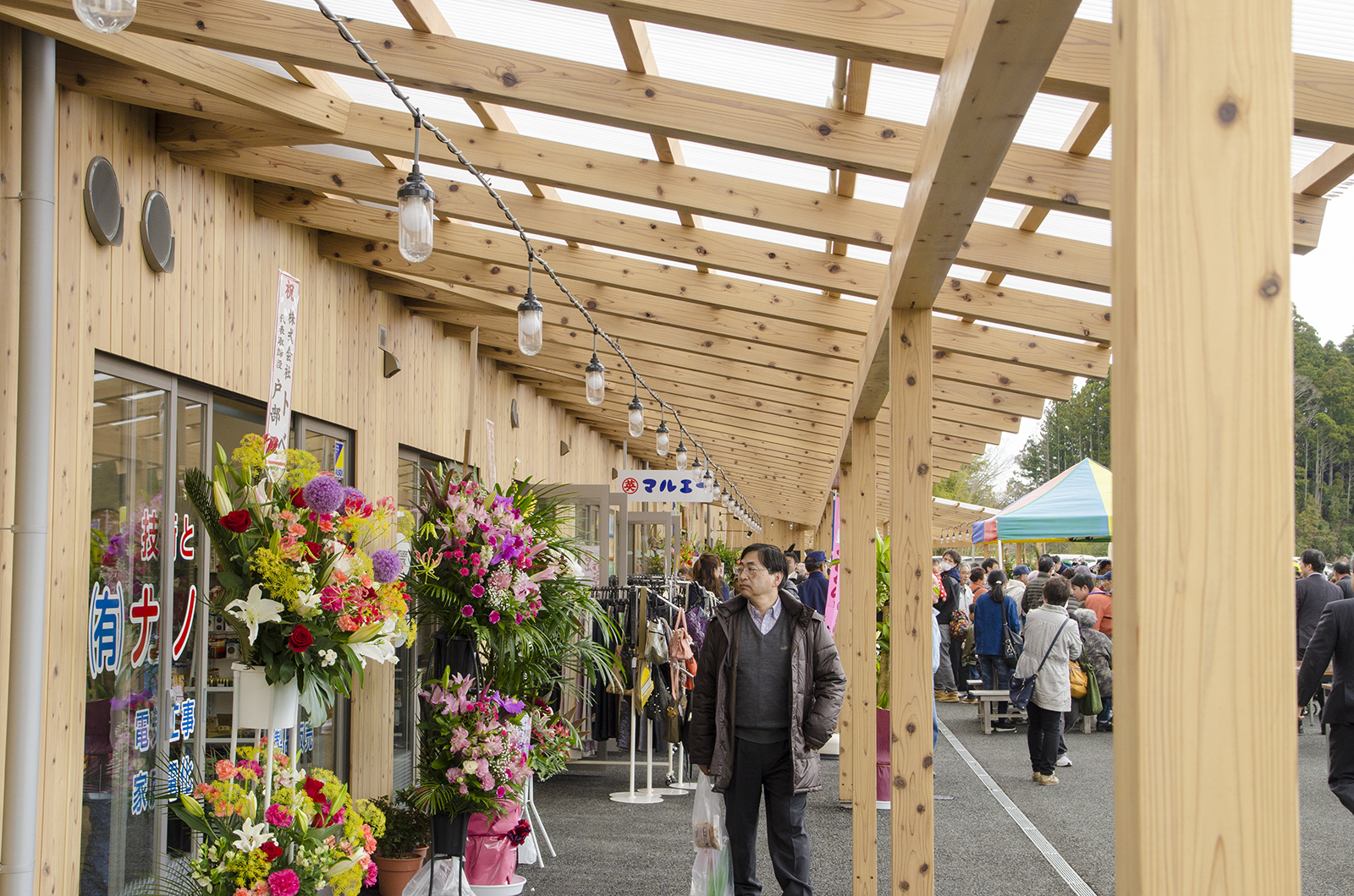 木造の軒下に開店祝いの花が並び多くの来場者が行き交う商店街の様子の写真