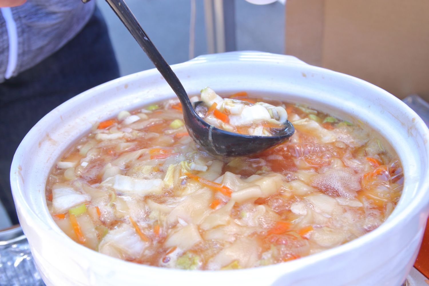 白菜や人参、ネギなどの野菜がたっぷり入った煮ぼうとうが煮え立っている大きな土鍋の中の具を、おたまですくい上げている写真
