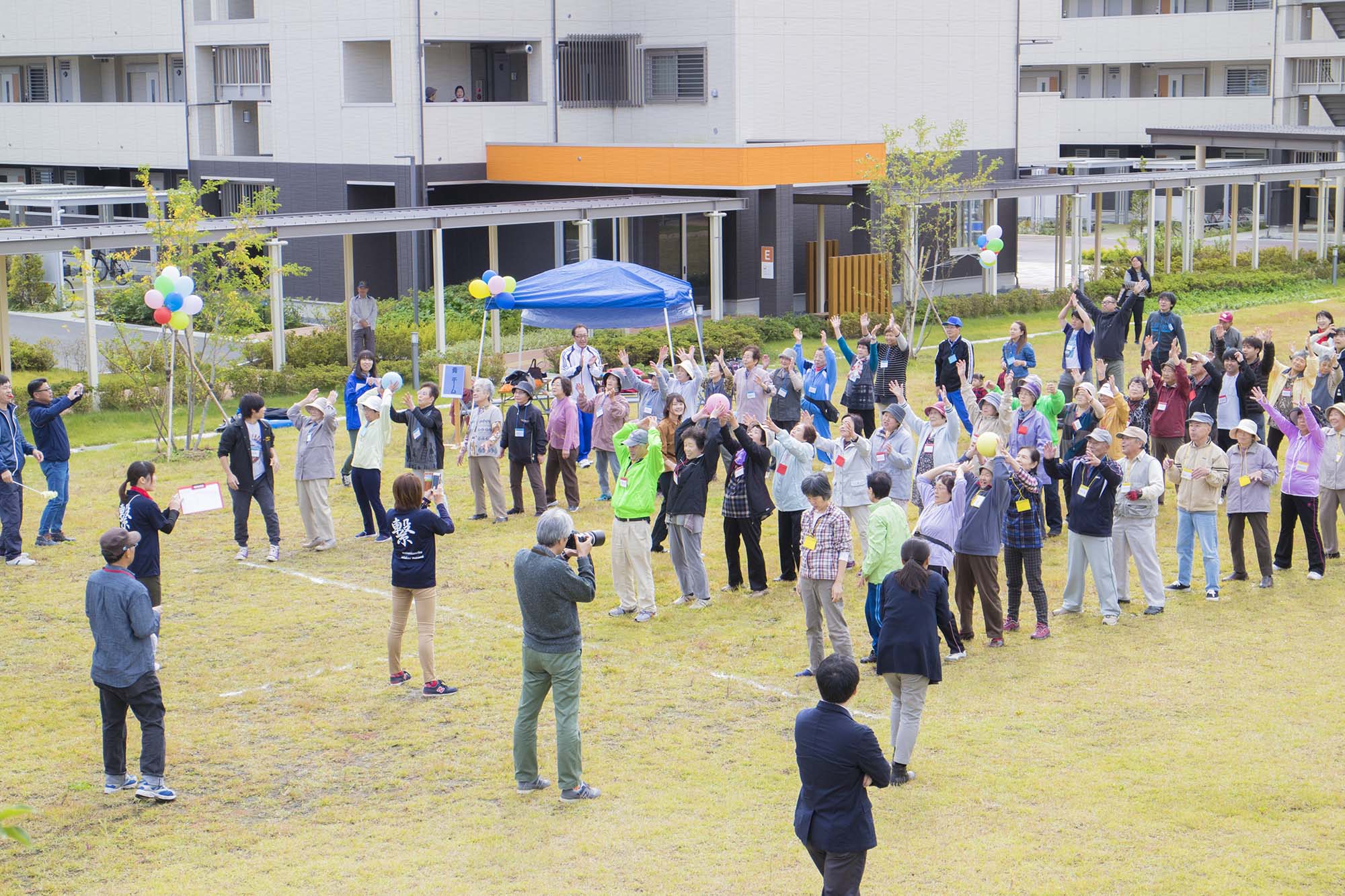 屋外の広場で多くの高齢者や地域住民が一斉に手を挙げたり身体を動かしたりして運動会の準備体操をしており、周囲にはカメラを持ったスタッフがカメラを構えている写真