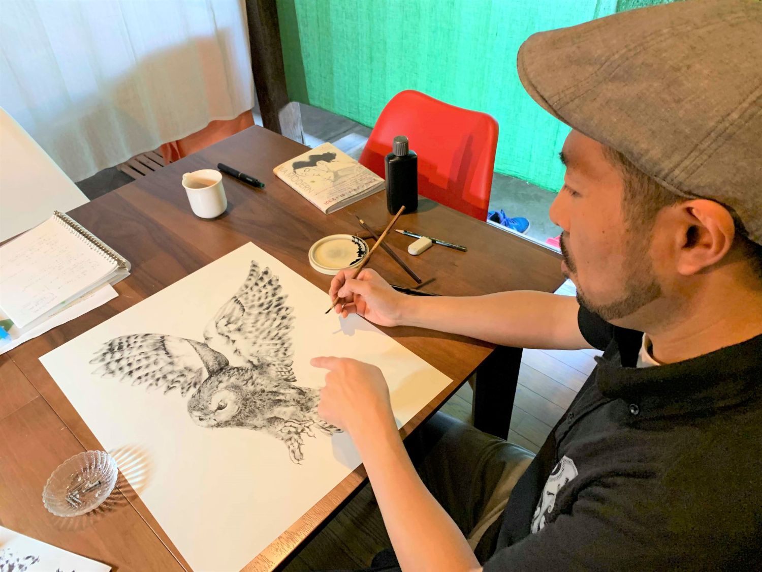 白いマグカップやスケッチブック、墨皿などが散らばっている机の上で、翼を広げたフクロウの緻密な墨絵を描いている男性の写真
