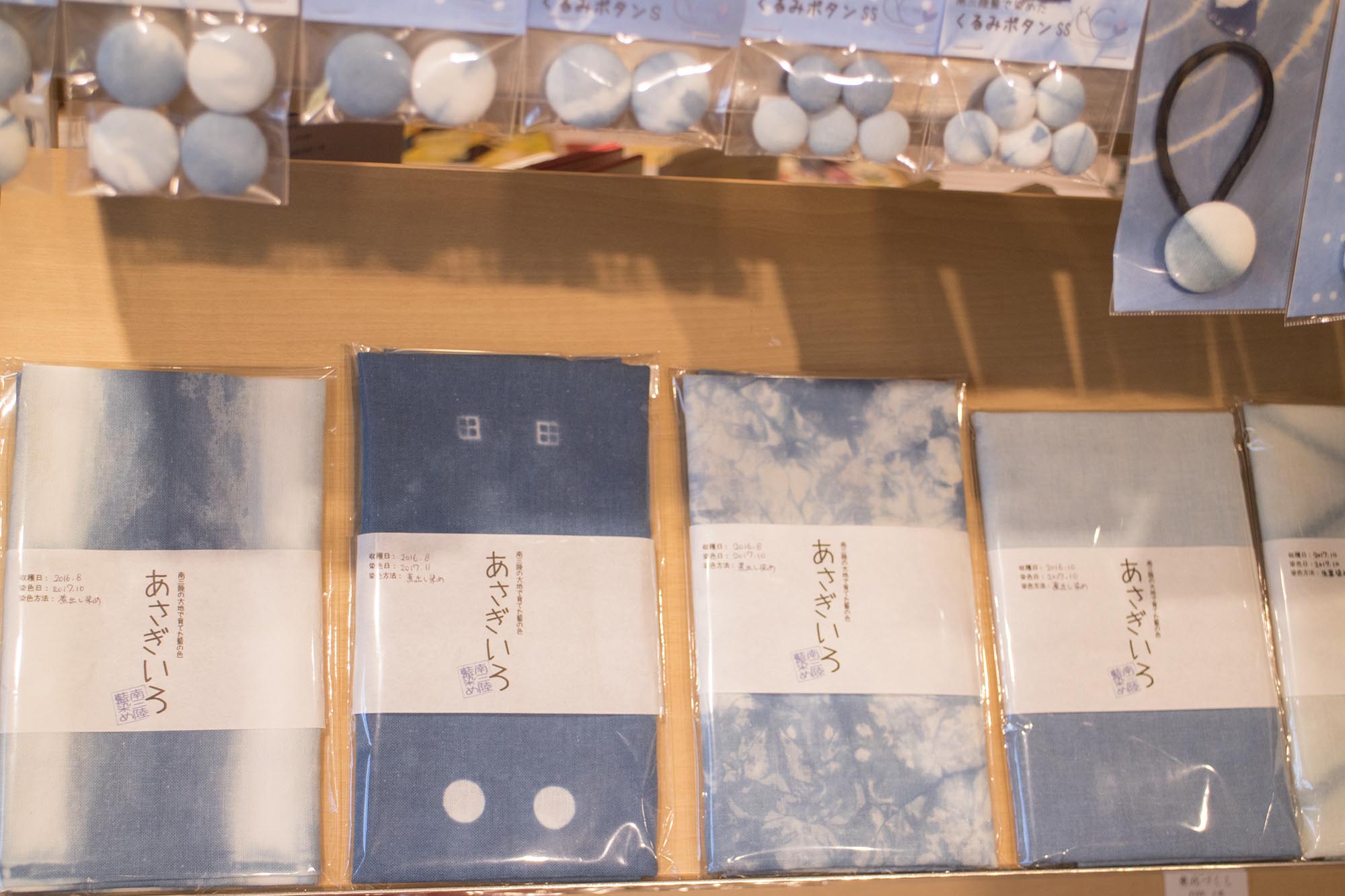 「あさぎいろ」と書かれたラベルが貼られた藍染の布製品が並び、その上に同じ色合いのくるみボタンなどが並んでいる写真