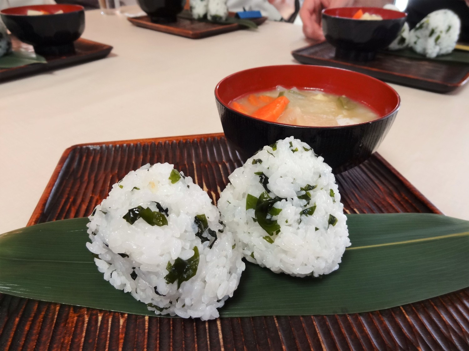 木製のトレイの上に、白いご飯で握られたワカメ入りのおにぎりが二つ並び、その横に赤い器に入った味噌汁が添えられている写真