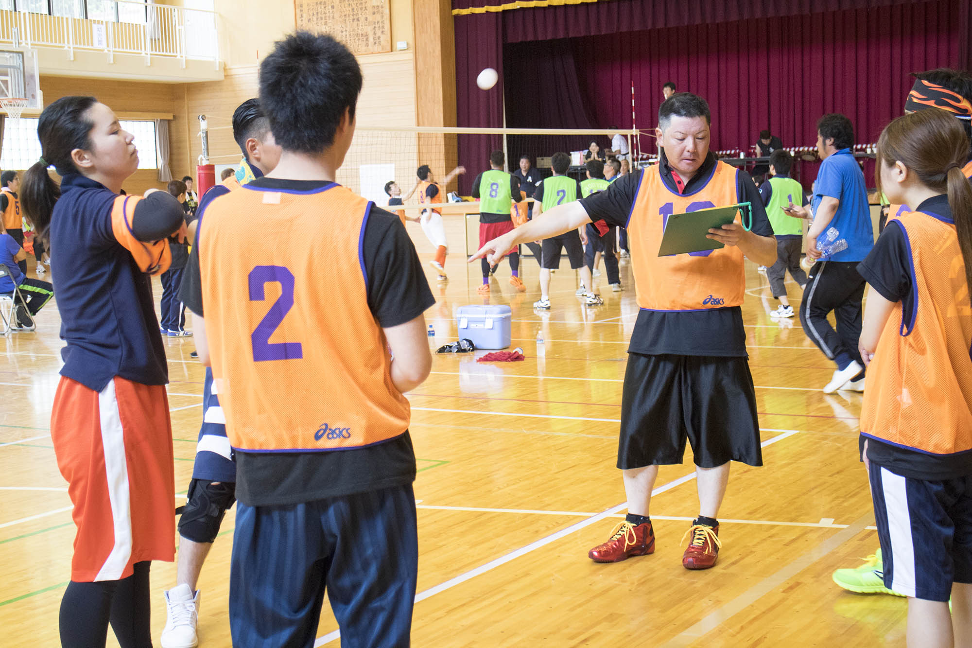 体育館で、オレンジのビブスを着たチームの1人の男性がボードを見ながら、選手たちに説明している写真