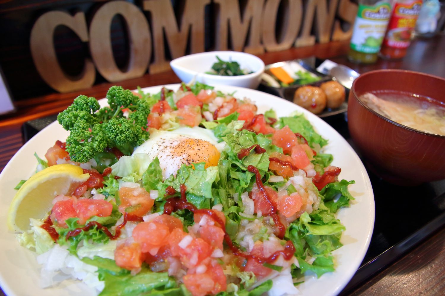 「COMMONS」の看板の前に置かれたトレイに置かれた、目玉焼きがのったたっぷりのレタスとトマト、タレがかかったタコライス、味噌汁、小鉢が添えられた看板メニューの写真