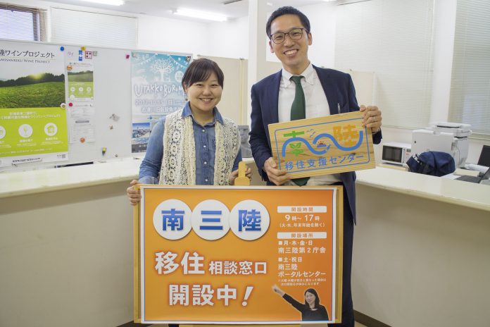 女性とスーツ姿の片山さんが室内で並んで立ち、「南三陸 移住相談窓口 開設中！」と書かれた大きなオレンジ色の看板と、「南三陸移住支援センター」と書かれた木製プレートを手に持っている写真