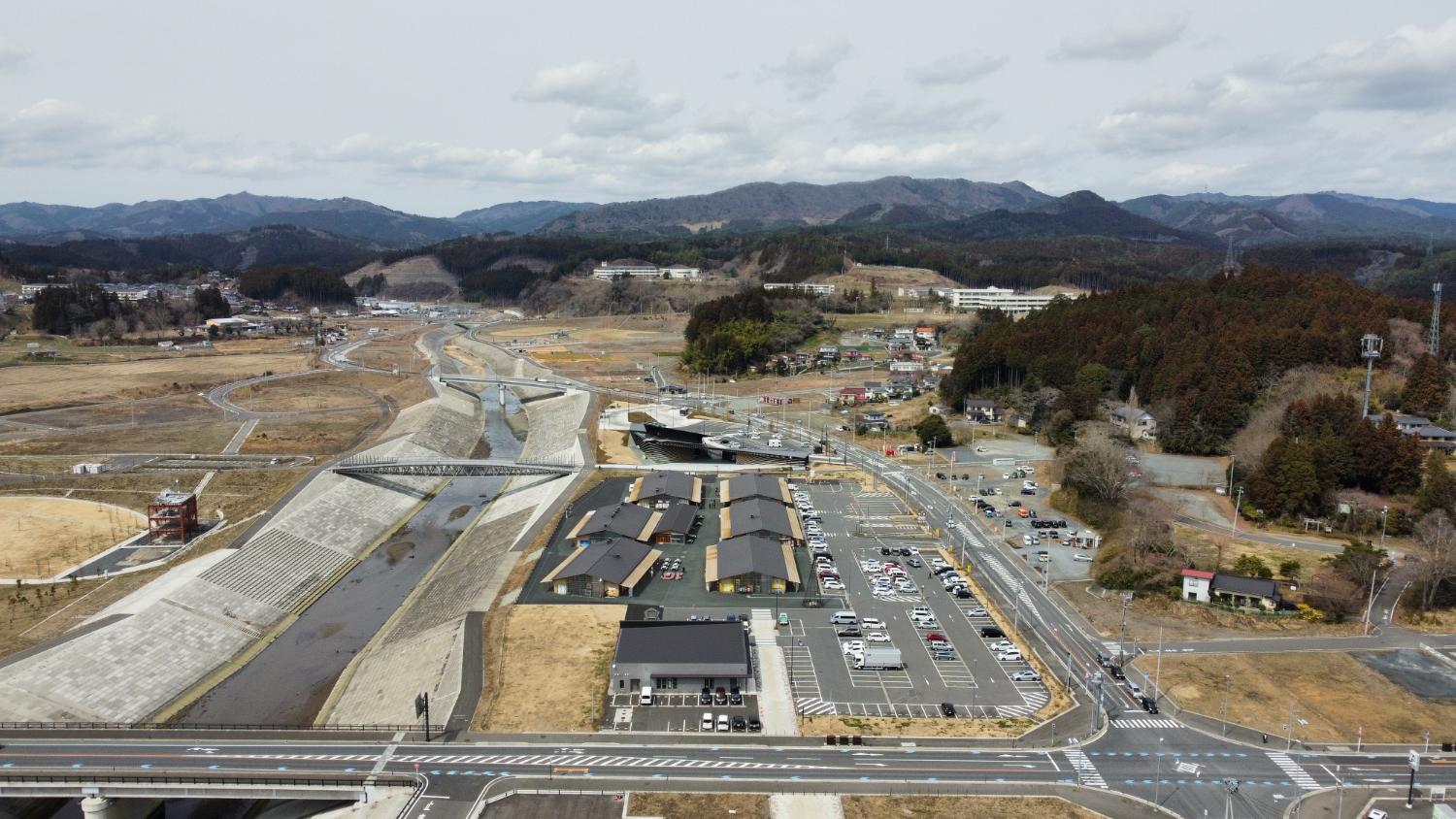 川沿いに整備された遊歩道と橋、中央に木造風の建物群と駐車場が広がり、その奥に住宅と学校らしき建物がある昼間の風景を高台から定点カメラで撮影した写真