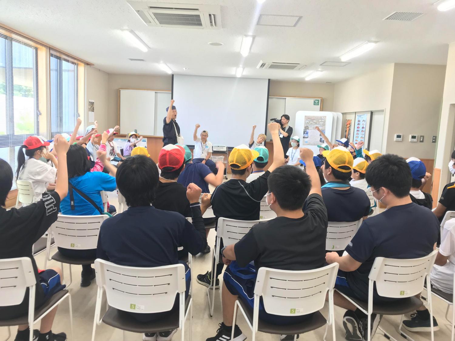 色とりどりの帽子をかぶった子どもたちが室内で椅子に座り、前に立つ先生に手を挙げながら元気に参加している写真