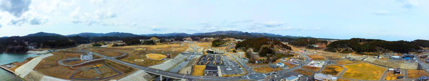 広大な更地に整備された道路網が広がり、背景には山並みが連なり、右手前に駐車場と建物が見える志津川市街地全景の昼間の風景を高台から定点カメラで撮影した写真