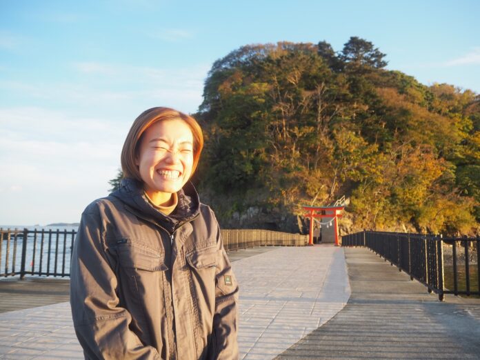 海辺の遊歩道に立つ渡邉陽子さんが、満面の笑顔で目を細めている写真