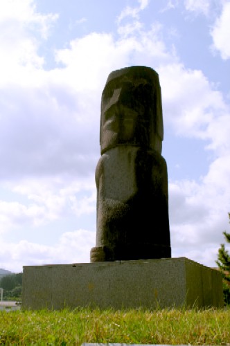 青空と白い雲を背景に、芝生の上に据えられた四角い台座の上に立つ巨大なモアイ像のレプリカを、下から見上げるように撮影した写真