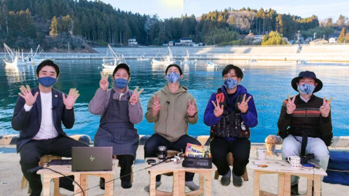 前のテーブルにはノートパソコンや釣り竿、飲み物が置かれている5人の男性が港のバーチャルセットを背景に並んで座り、全員がマスクをしてカメラに向かって両手でピースサインをしている写真