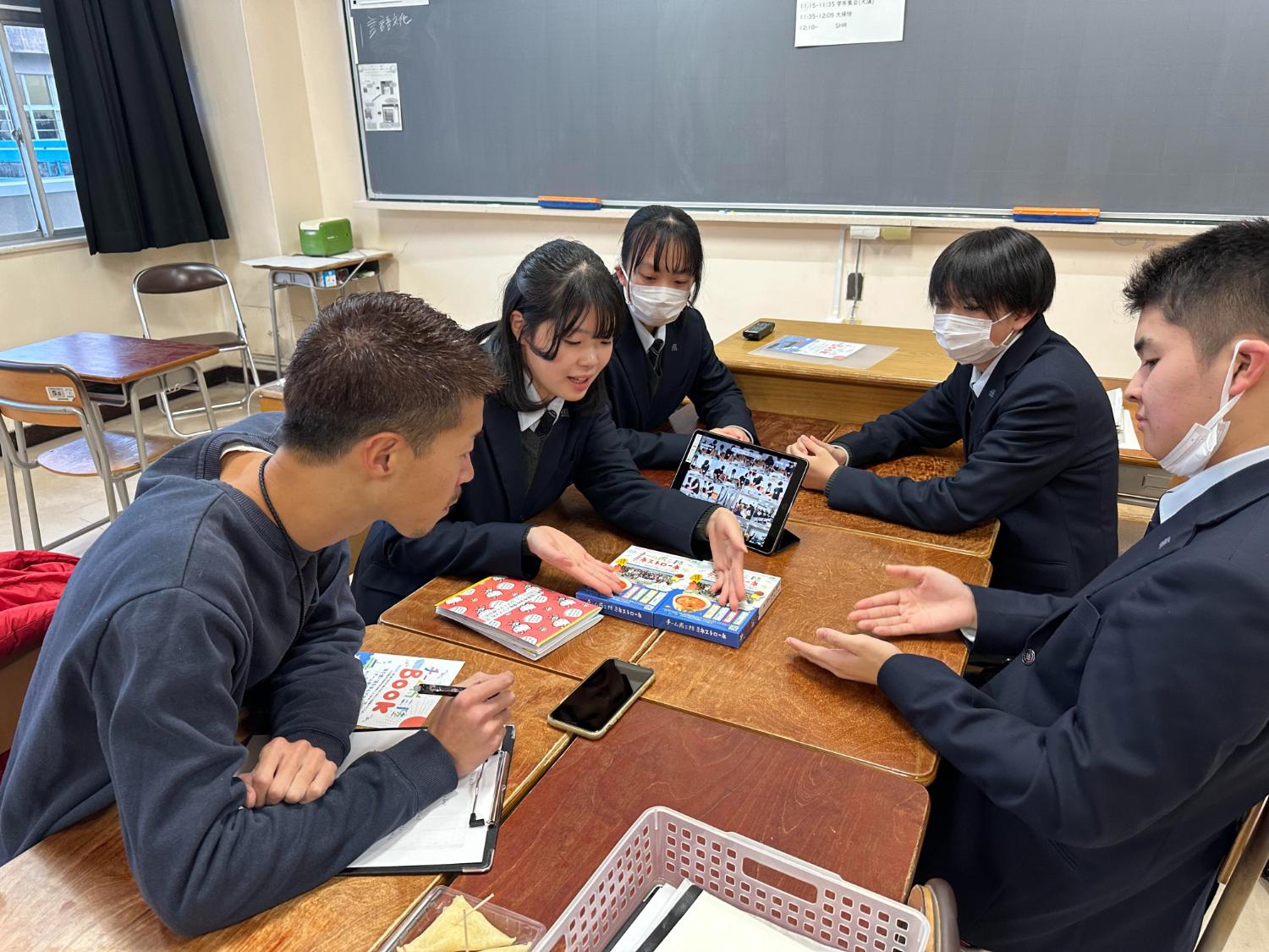 南三陸高校の生徒たちが教室で冊子やタブレットを囲み、南三陸ミネストローネの魅力をどう伝えるか話し合っている写真