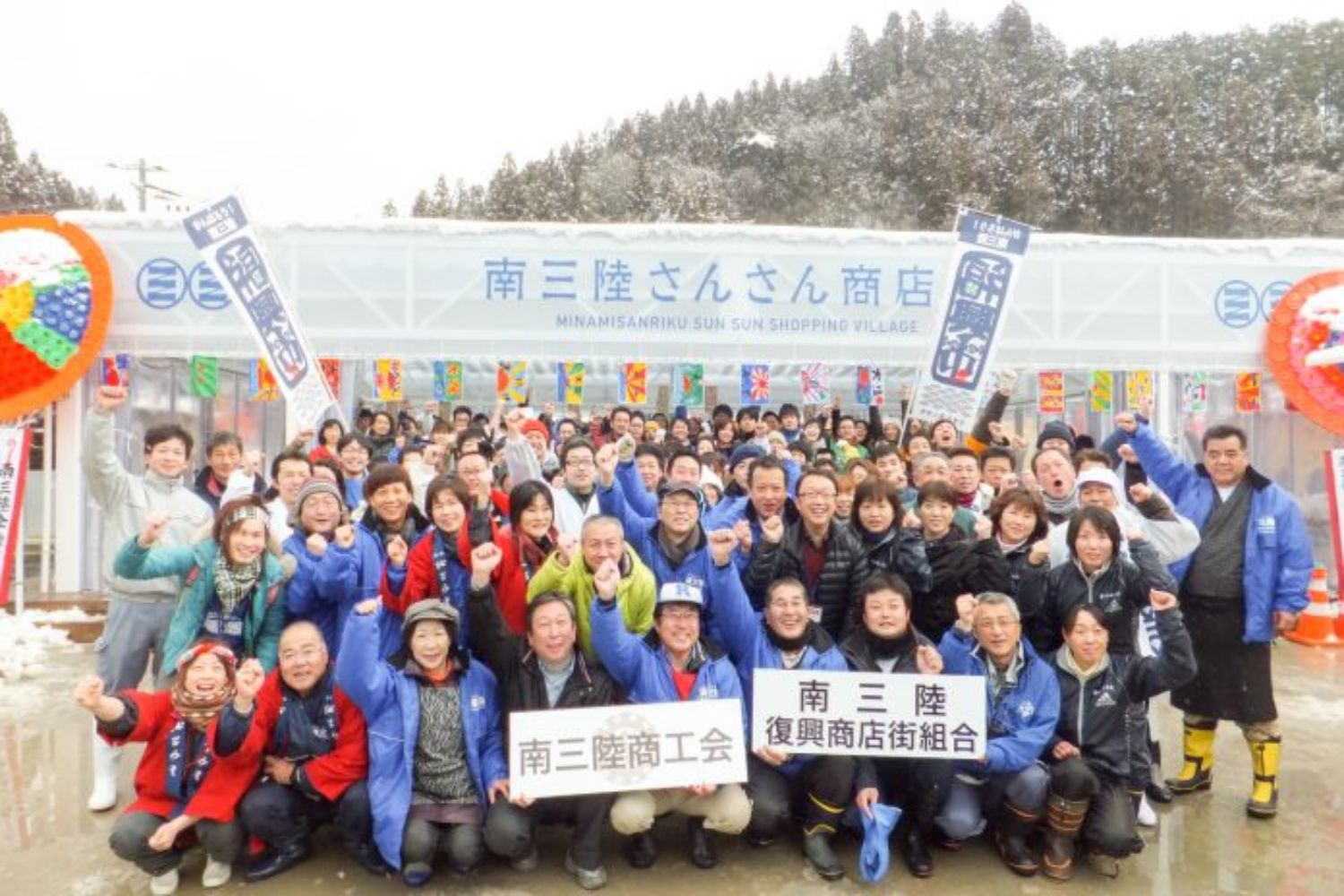 雪が残る中、「南三陸さんさん商店街」の前で大勢の人々が集まり、「南三陸商工会」「南三陸復興商店組合」と書かれたボードを持ち、笑顔でガッツポーズをしている仲間たちの集合写真