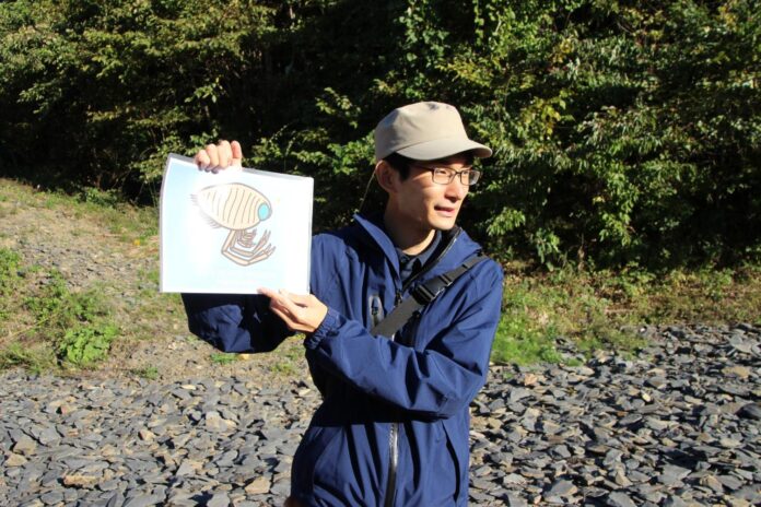 帽子をかぶりメガネをかけた菊池優さんが左手で昆虫のイラストが描かれた紙を持ち説明する様子の写真