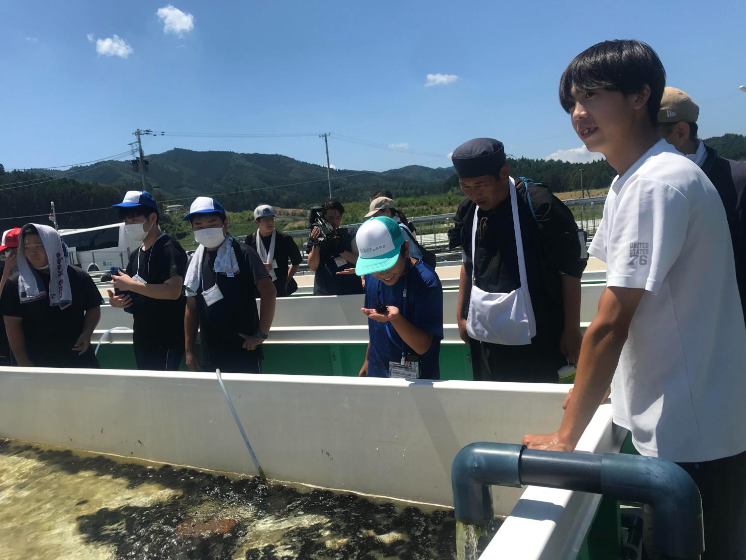 屋外の養殖施設で高校生と子どもたちが水槽をのぞき込み、南三陸の海産物について学んでいる写真