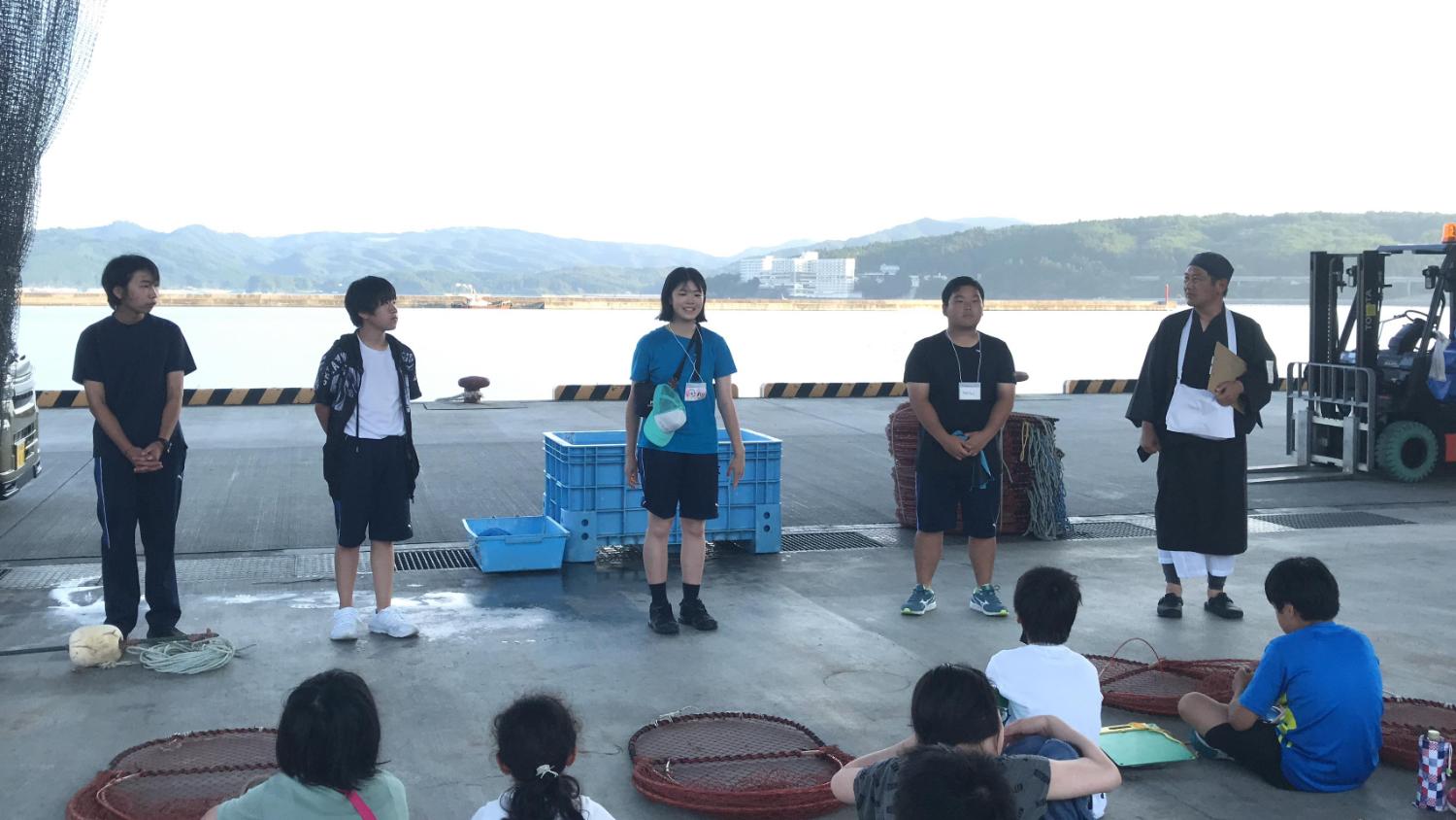 港の岸壁で南三陸高校の生徒と漁師が並び、海について学ぶために座って話を聞く子どもたちの前で説明している写真