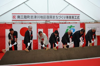 「南三陸町志津川地区復興まちづくり事業着工式」と書かれた垂れ幕の下で、スーツ姿の男性たちが並んで鍬入れをしている写真