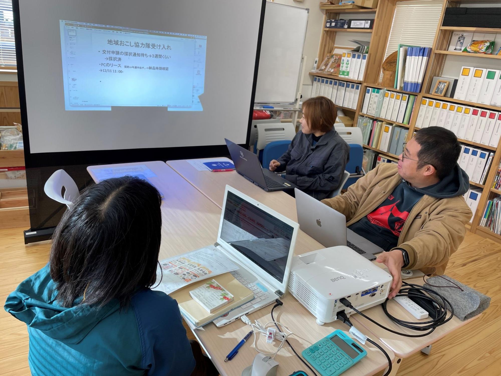 会議室で3人がプロジェクターを使い、スライドを見ながら話し合っている様子の写真