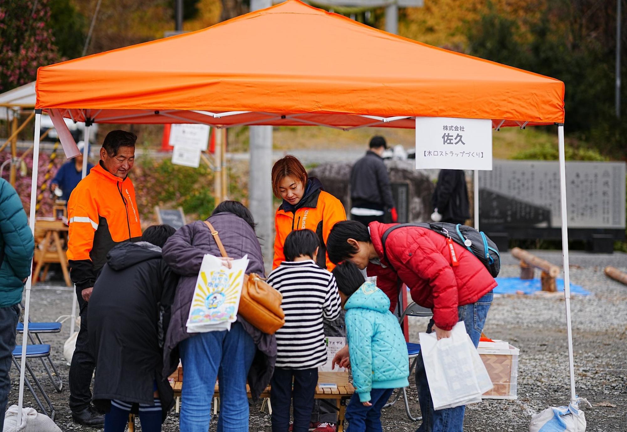 屋外のオレンジ色のテントの下で、大人と子どもが集まり何かを見たり触ったりしている様子の写真