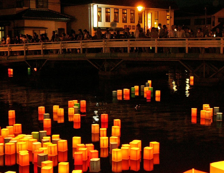 夜の八幡川の水面に多数の灯籠が浮かび、温かい光を放ちながら流れていく様子と、橋の上に集まった多くの人々がそれを見守っている流灯会の写真
