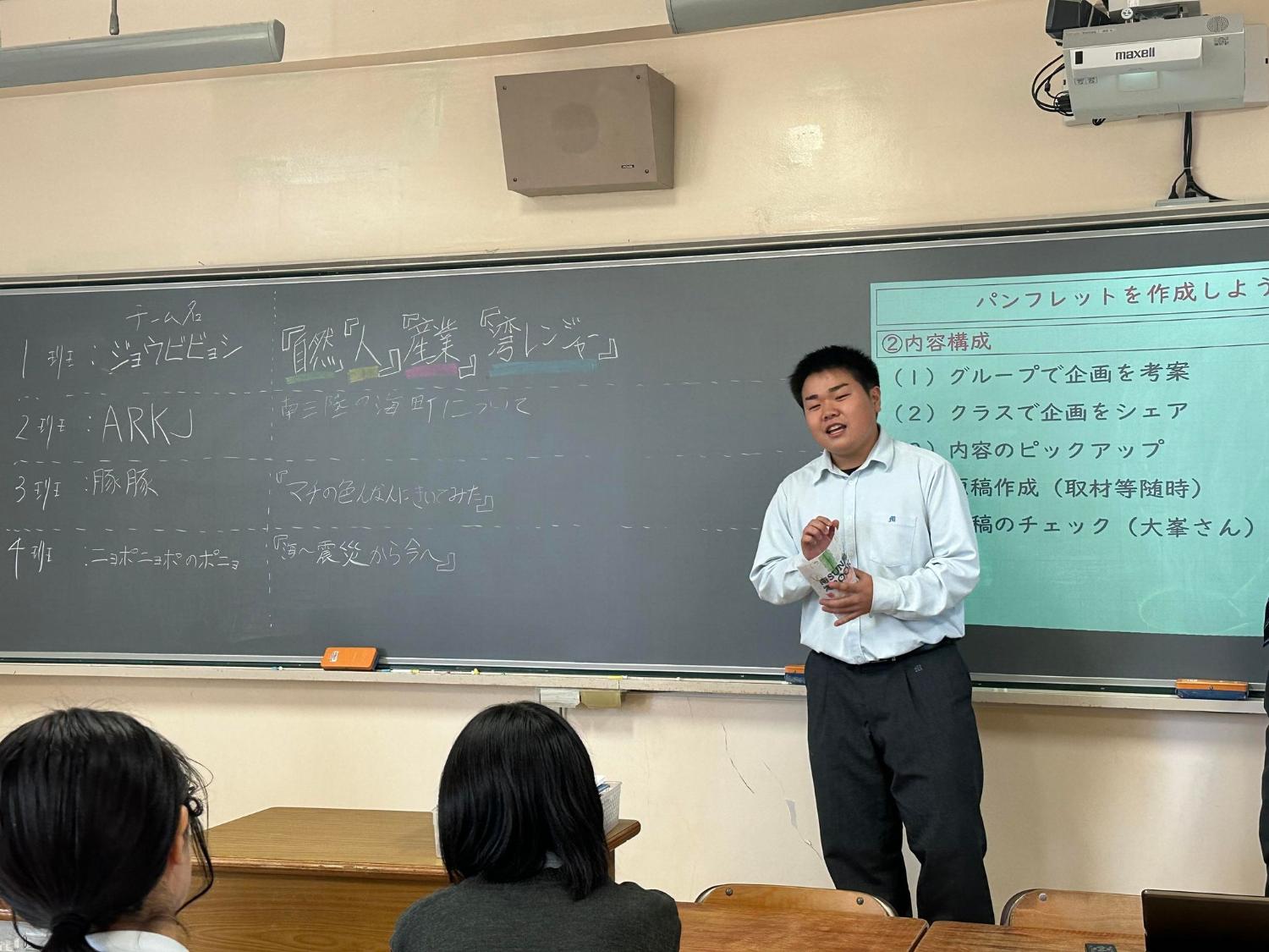 教室で一人の高校生が黒板の前に立ち、グループで考えたパンフレットの企画内容をクラス全体に発表している写真