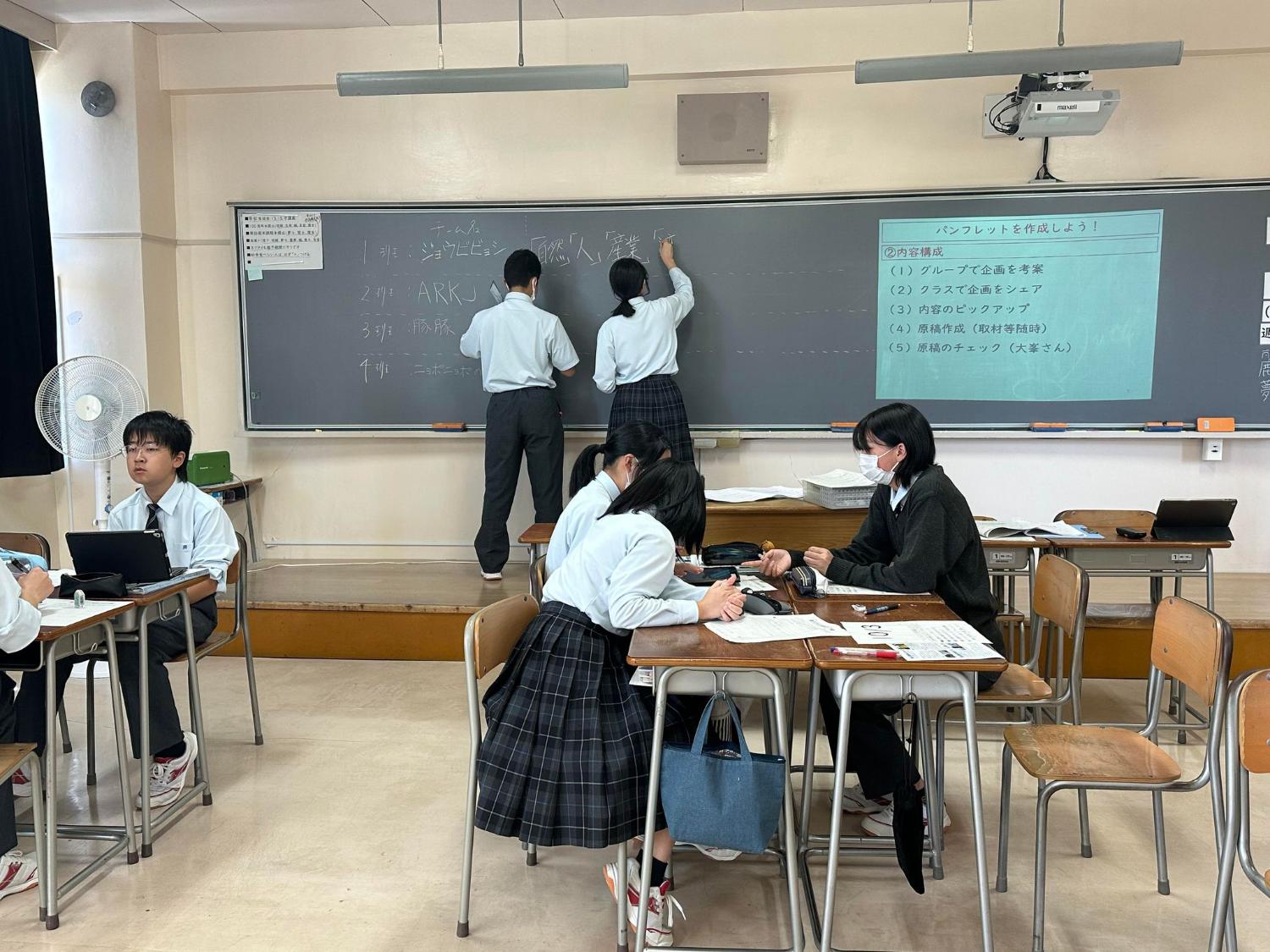 黒板の前で高校生がグループに分かれて座り、パンフレット制作に向けて企画や構成を話し合っている写真