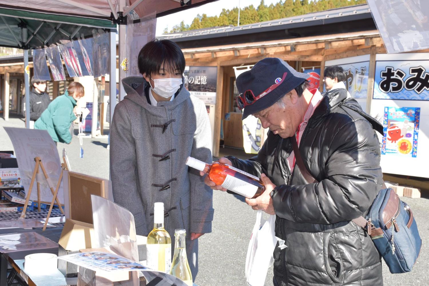 テントで、高校生が南三陸産の商品について来場者に説明し、来場者が瓶詰めの商品を手に取って確認している写真