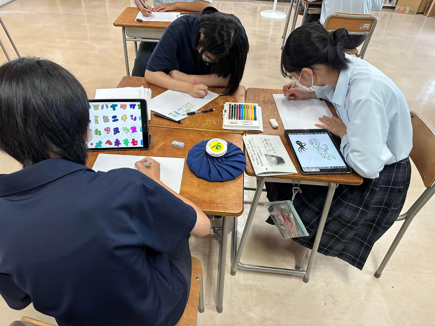 高校生が教室の机でカラーペンやタブレットを使い、南三陸ミネストローネのキャラクターやイラストを描いている写真