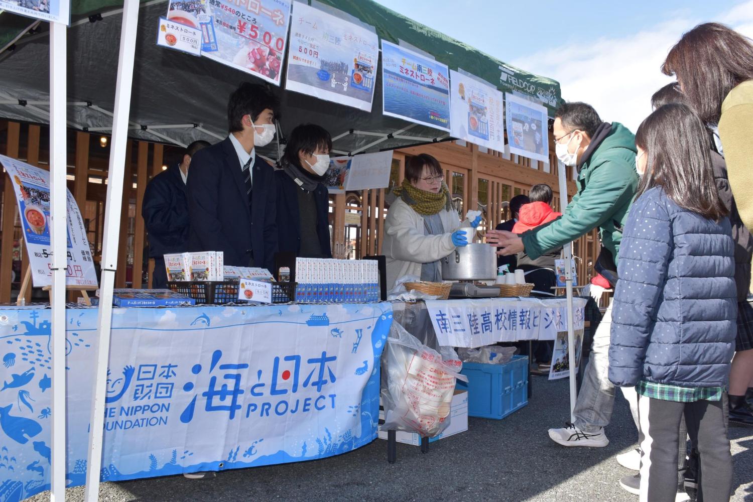 屋外のイベント会場で、高校生が南三陸ミネストローネを来場者に提供しながら、地域の魅力を紹介している写真