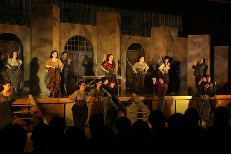 薄茶色の石造り風の立体的なセットの前で、中世ヨーロッパ風の衣装をまとった十数人のキャストが立ち、歌いながら感情豊かに演技を披露しているオペラ公演の様子を写した写真