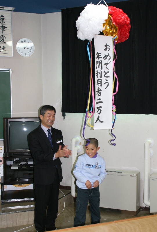 くす玉が割られ、『おめでとう 年間利用者2万人』の垂れ幕が下がっており、スーツ姿の男性が拍手しており、その横に立つ山内翔太さんが笑顔で喜んでいる様子の写真